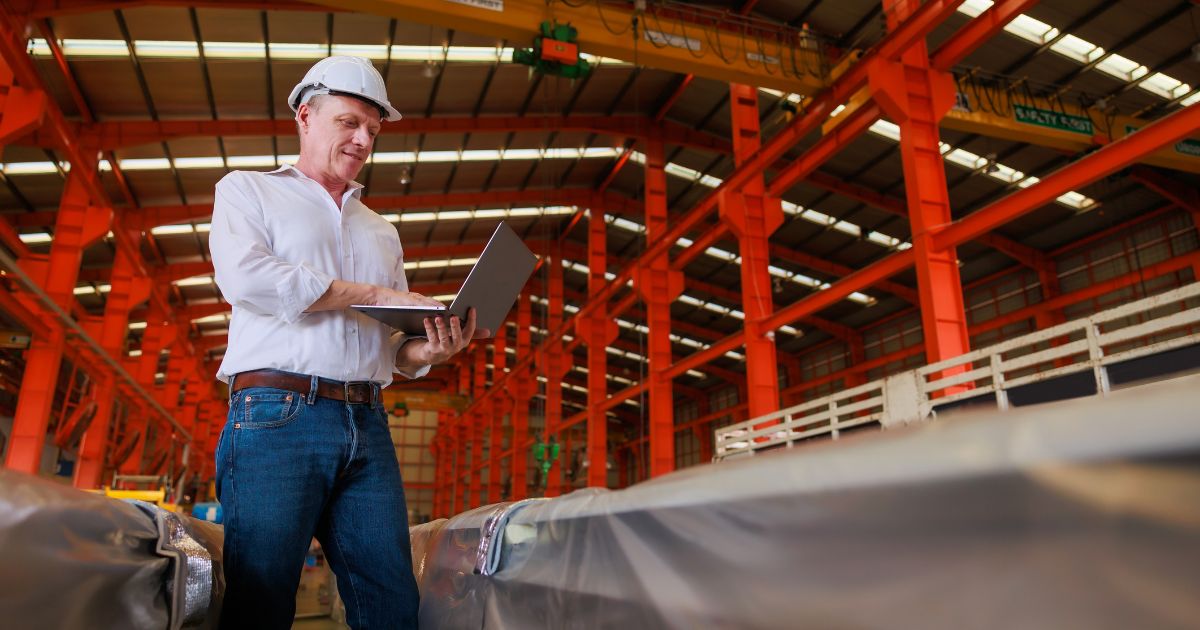 Man in an industrial warehouse wearing a hard hat and using a laptop.