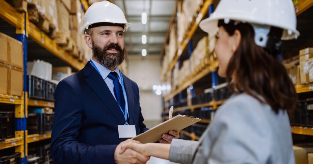 Woman and man in suits and hardhats shaking hands in a warehouse