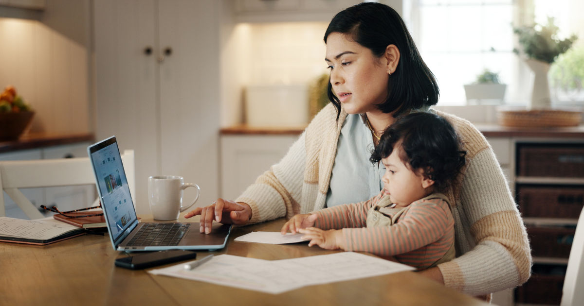 Woman holding baby at table reading news on laptop